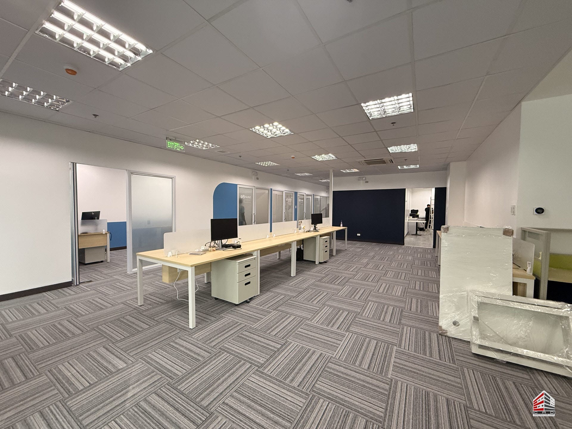 A wide-angle view of a modern open-plan office featuring rows of light wood workstations with white legs, grey striped carpet tiles, and integrated frosted glass privacy dividers.