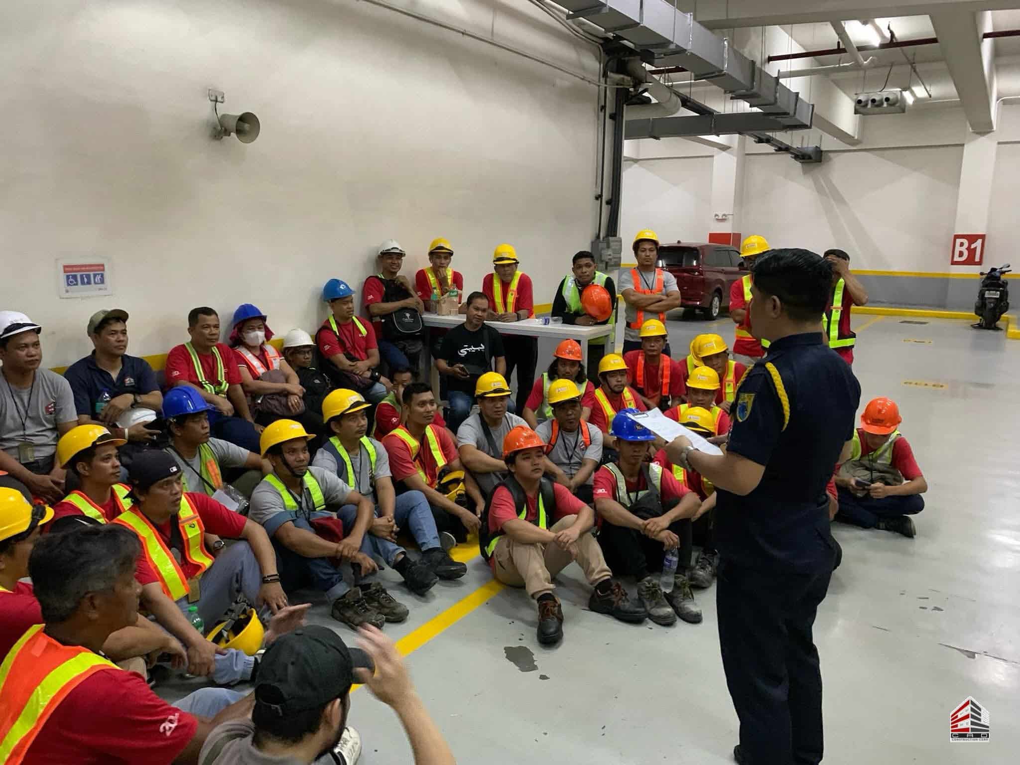 A safety officer in a dark blue uniform stands in the foreground holding a clipboard, addressing the group. The workers are wearing various colored hard hats and high-visibility safety vests.