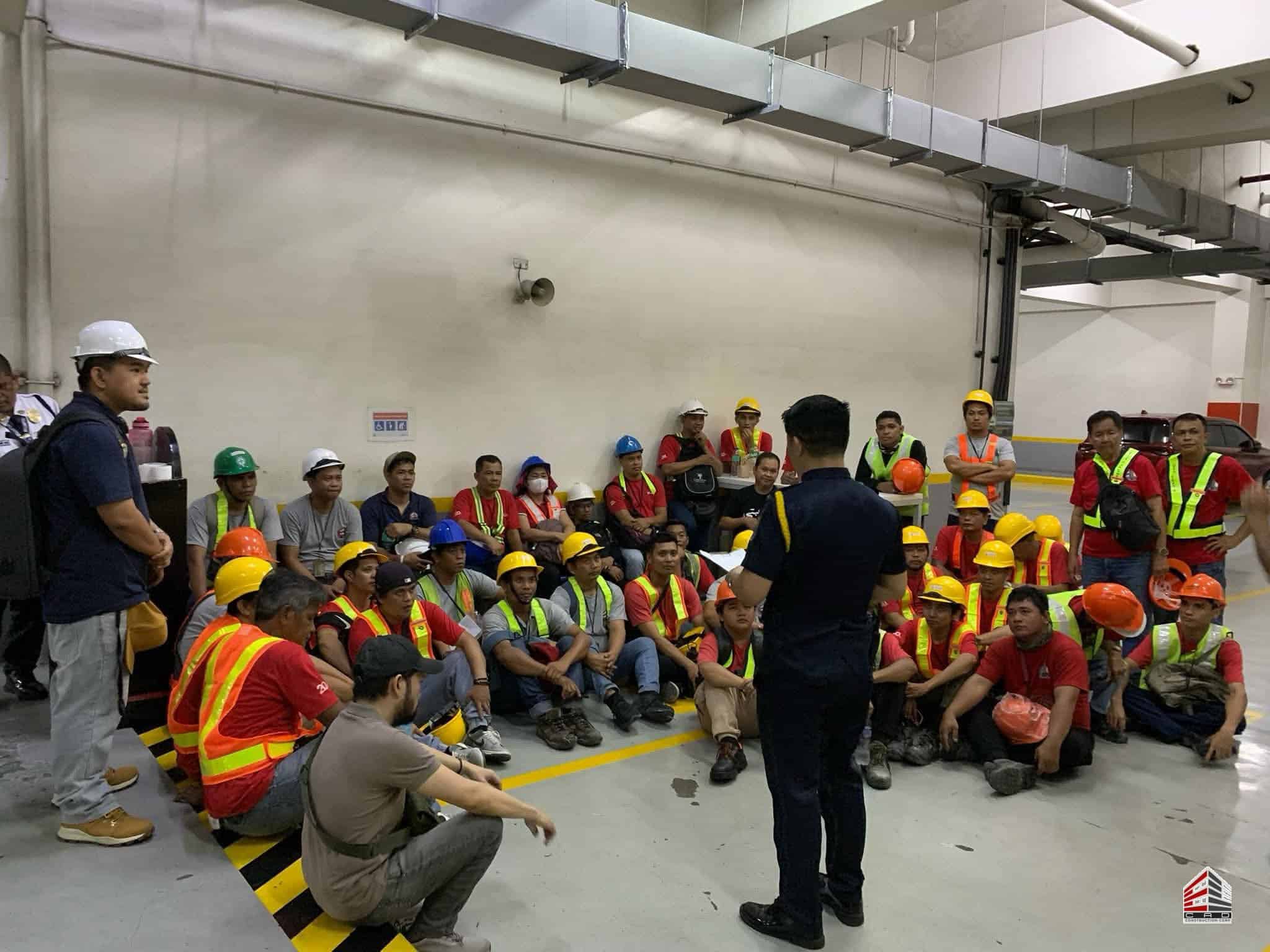 The construction team is gathered in a parking level, listening intently to the speaker. Several workers are in the foreground, and the composition highlights the organized group setting and compliance with safety protocols.