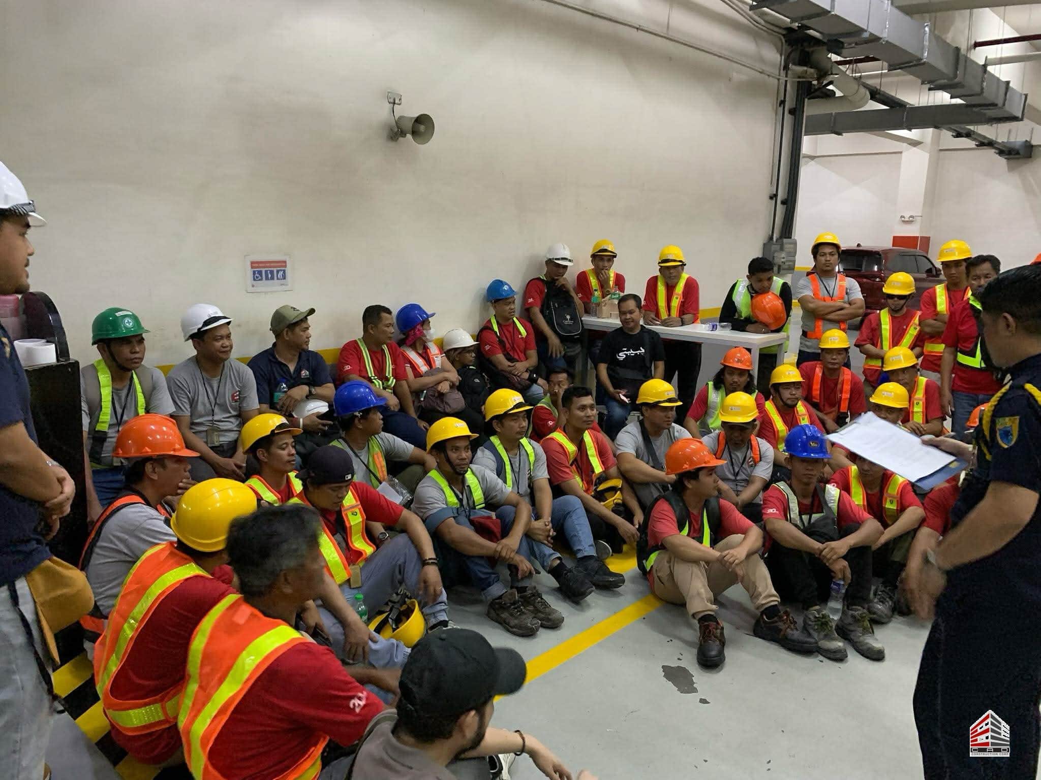 A close-up perspective of the construction workers seated during the orientation session. The focus is on the diverse group of personnel in their safety gear, with the safety officer visible on the right side of the frame holding briefing documents. The background shows the industrial setting of a building's lower level.