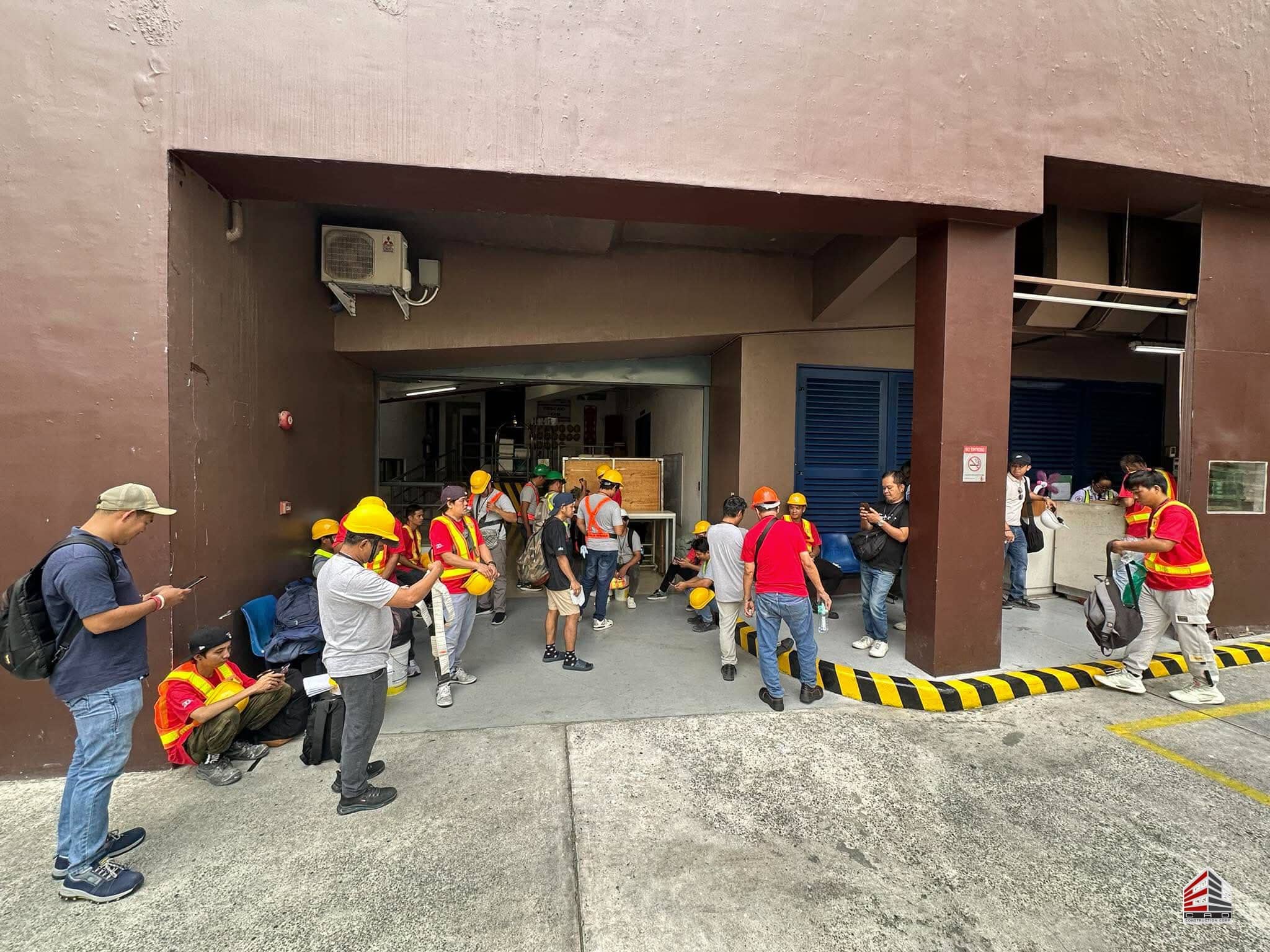 Construction personnel gathered at the shaded entrance of the project site, preparing for the day's tasks with their safety gear.
