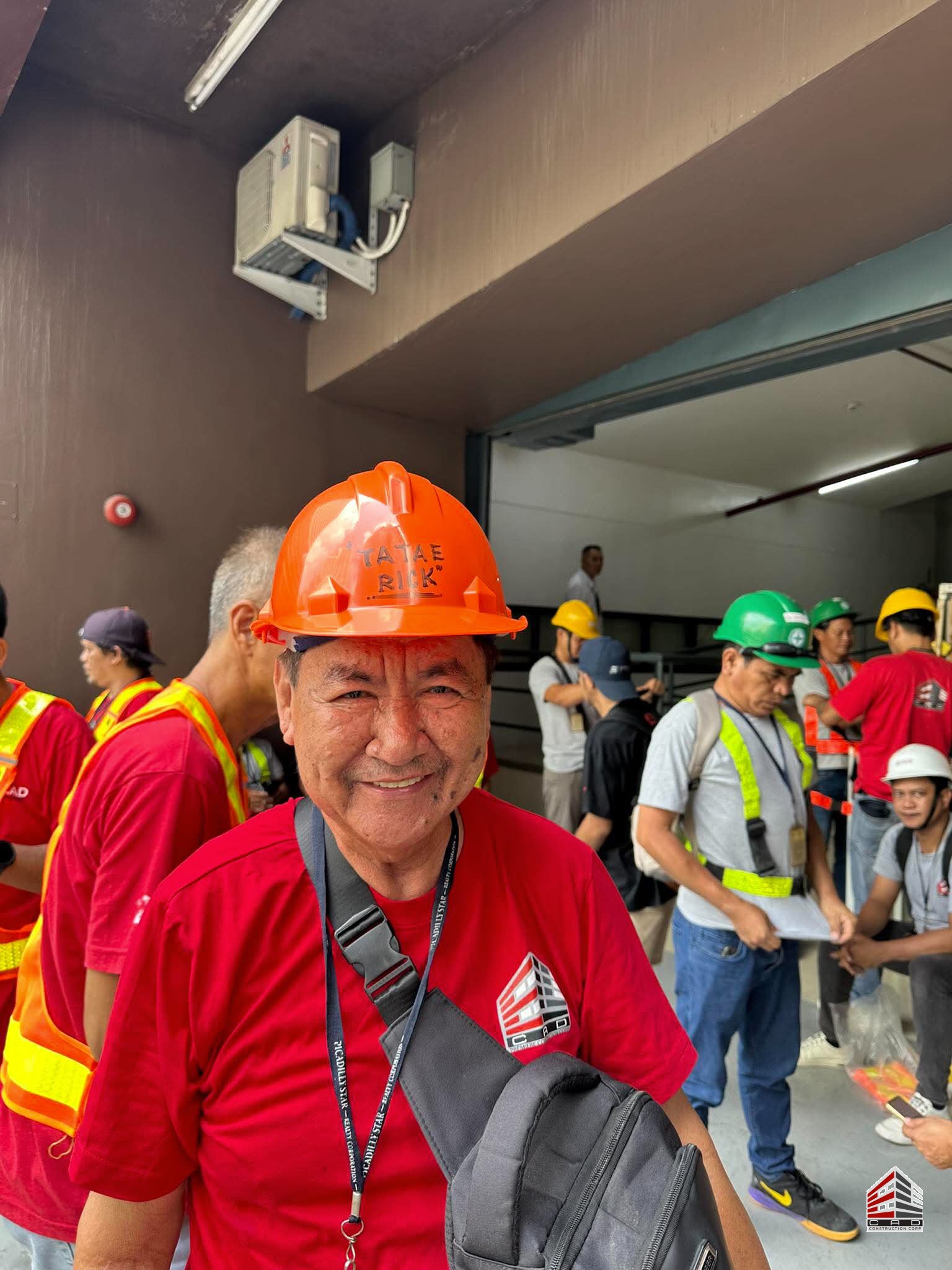 A portrait of a smiling construction worker in a red Ellcad shirt and orange hard hat, with colleagues in PPE visible in the background.